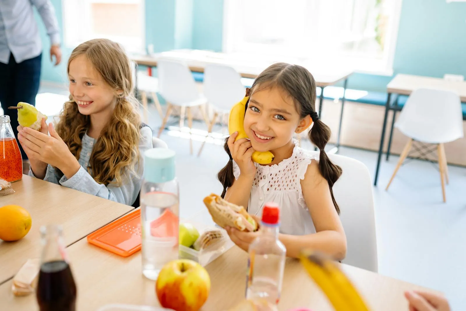 Two smiling schoolgirls eating fruit and a sandwich at lunch